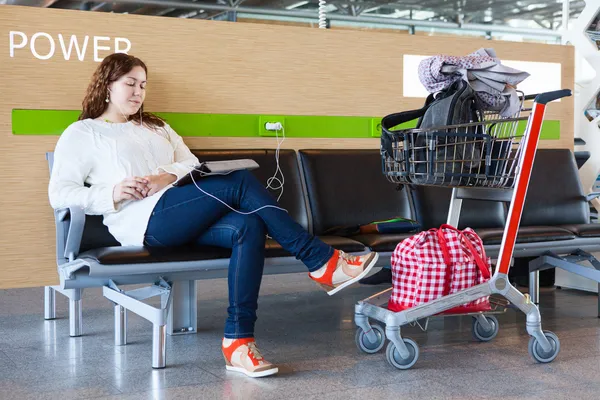 Tired woman charging tablet pc in airport lounge with luggage hand-cart