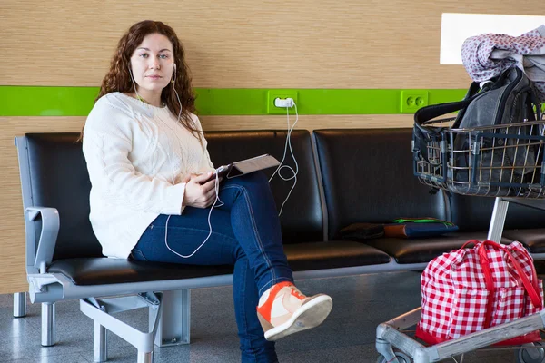 Serene woman charging tablet pc in airport lounge with luggage hand-cart