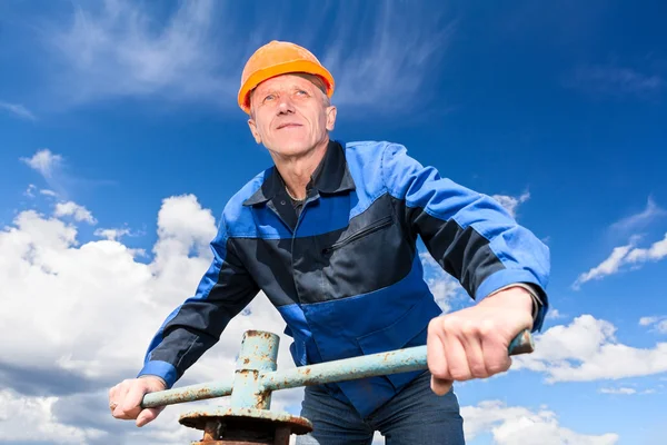 Senior Caucasian worker in a hardhat at the factory with the pipe valve against blue sky
