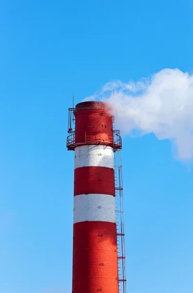 Red and white factory chimney with smoke against blue sky