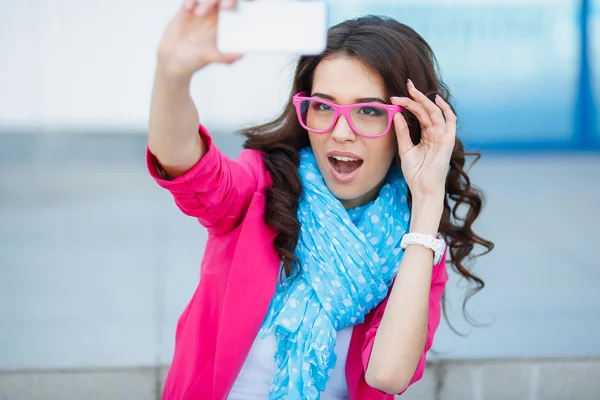 Happy young girl making funny face while taking pictures of herself through cellphone, over white background