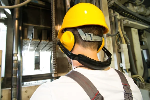 Worker in safety hat and headphones working behind machine on a factory