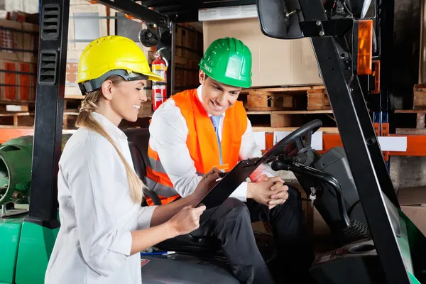 Supervisor Showing Clipboard To Colleague Sitting In Forklift