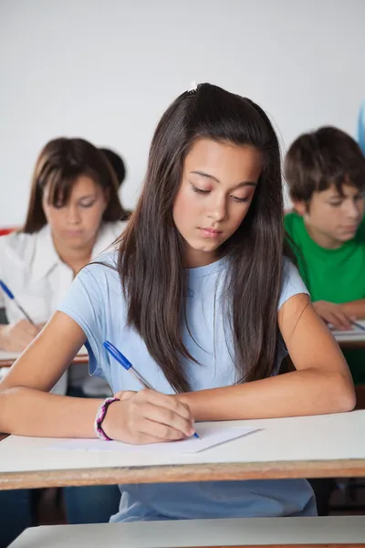 Female Student Writing Paper At Desk In Classroom