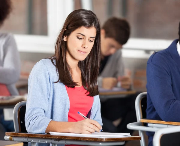 Woman Writing On Book At Desk In Classroom