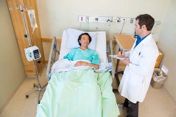 Male Doctor Looking At Patient While Writing Notes On Clipboard