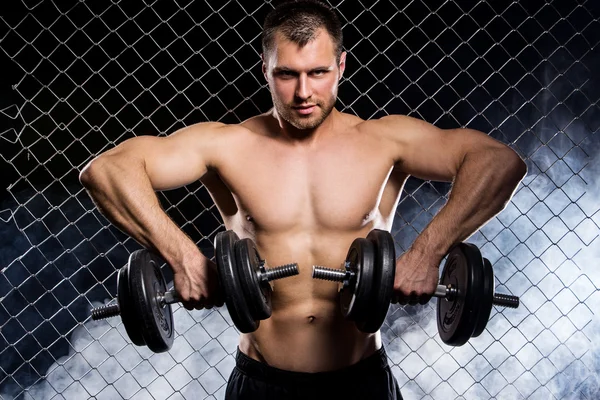 Powerful guy with a dumbbells showing muscles on fence backgroun