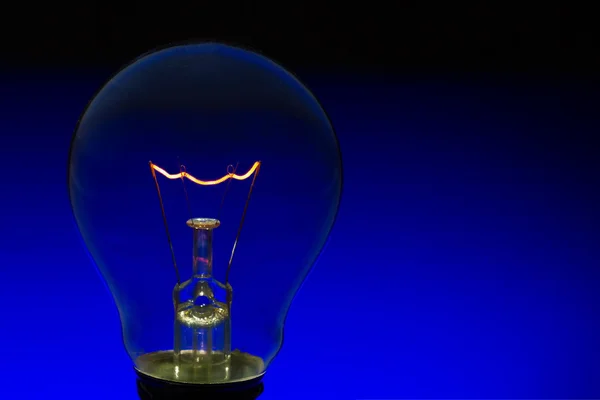 Glass light bulb with burning filament upright with blue backgro