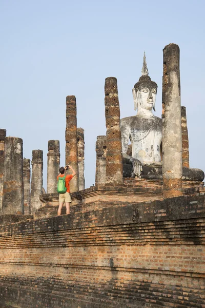 Man-tourist photographing the seated Buddha on the ruins of an ancient temple. The Sukhothai, Thailand