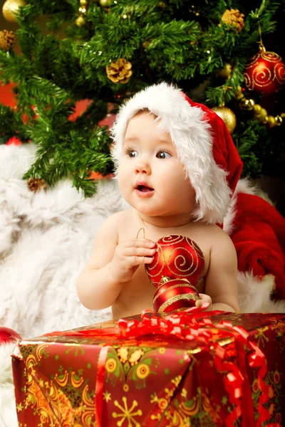 Christmas baby in hat holding red ball near gift box
