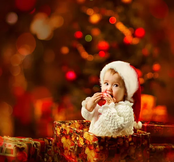 Christmas baby in Santa hat holding red ball near present gift box