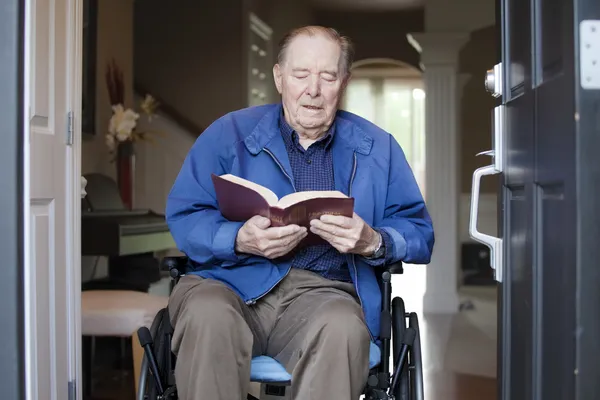 Elderly man in wheelchair at his front door reading the Bible