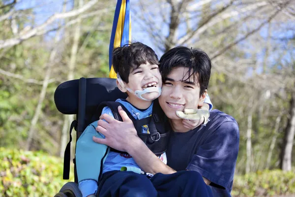 Little disabled boy in wheelchair hugging older brother outdoors