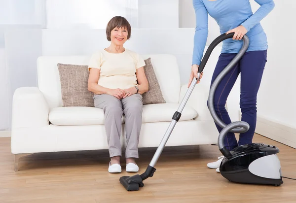 Caretaker Cleaning Floor While Senior Woman Sitting On Sofa