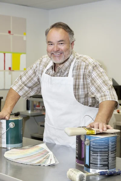 Portrait of a cheerful mature store clerk with paint cans and brush in hardware shop