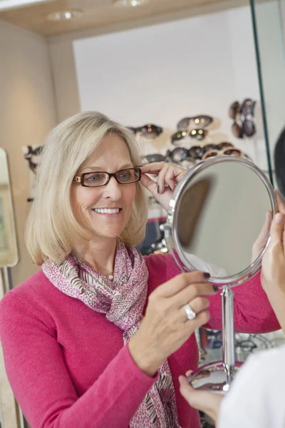 Happy senior woman trying on glasses while optician holding hand mirror