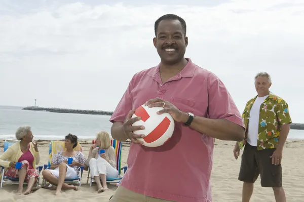 Man Holding Volley Ball At Beach With Friends Grouped Behind