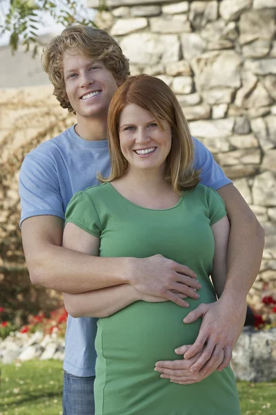 Pregnant Woman With Man Embracing From Behind Outdoors