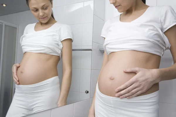 Pregnant woman stands looking at stomach in bathroom mirror