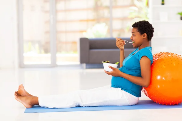 Pregnant african american woman eating healthy salad on exercise mat