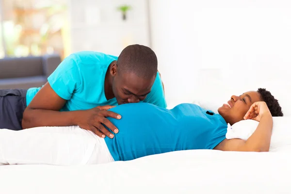 Young african american man kissing pregnant wife\'s belly on bed