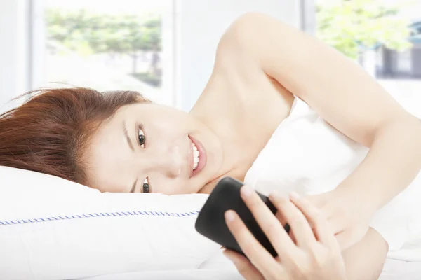 Smiling young woman texting by smart phone on the bed