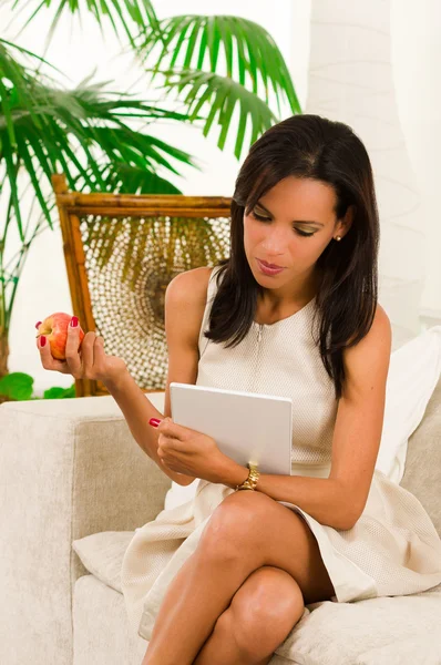 Beautiful young elegant woman sitting in livingroom using digital tablet and eating an apple