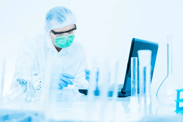 Scientist working in the lab, in protective mask and cap, examines a test tube with liquid