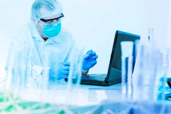 Scientist working in the lab, in protective mask and cap, examines a test tube with liquid