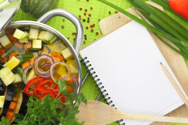 Blank recipe book with vegetable soup, kitchen equipment and veggies around them