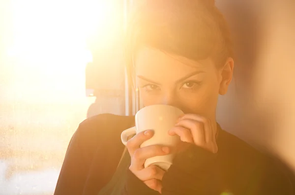 Beautiful young woman with cup of hot coffee over sun window
