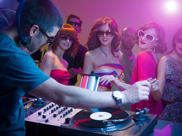 Male dj playing music with turntables and headphones at a party with dacing in front of him wearing sunglasses, with other in background and colorful lights
