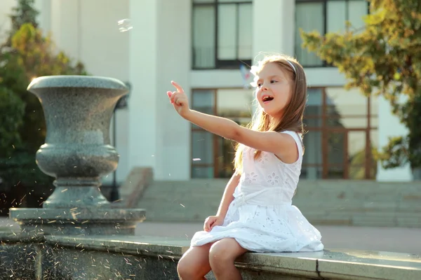 Cute smiling young girl in a beautiful white dress flounder feet in the fountain and blowing soap bubbles