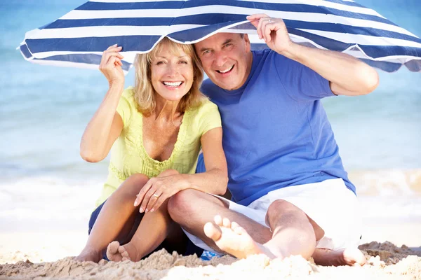 Senior Couple Sheltering From Sun Under Beach Umbrella