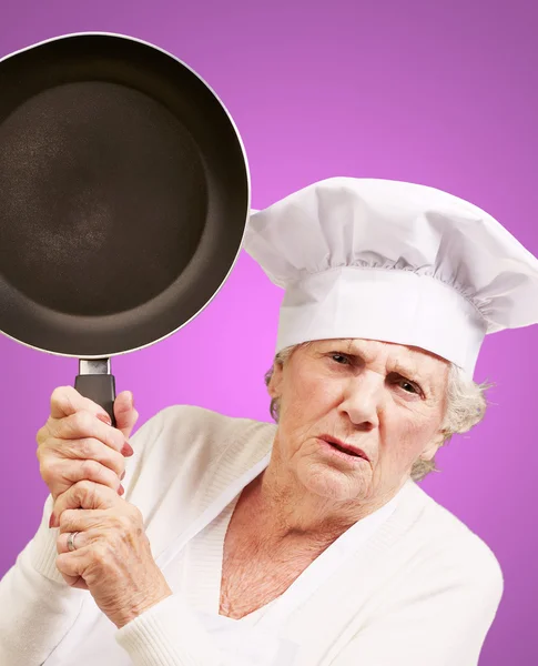 Cook senior woman angry trying to hit with pan over purple backg