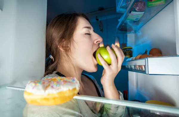 Portrait from inside of fridge of young woman eating apple