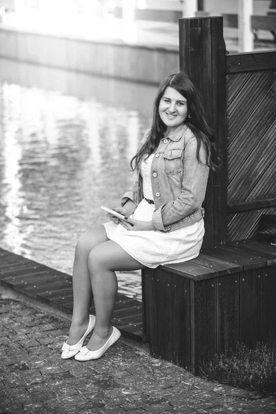 Young woman sitting on bench near river with tablet