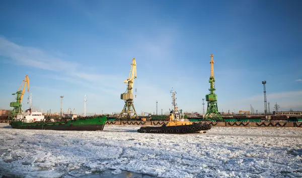 Small tug boat goes with vessel through icy channel in harbor of St.Petersburg cargo port