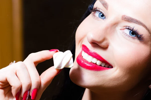 Closeup portrait on seductive charming beautiful young woman with blue eyes, red lips & hand with red nails holding candy