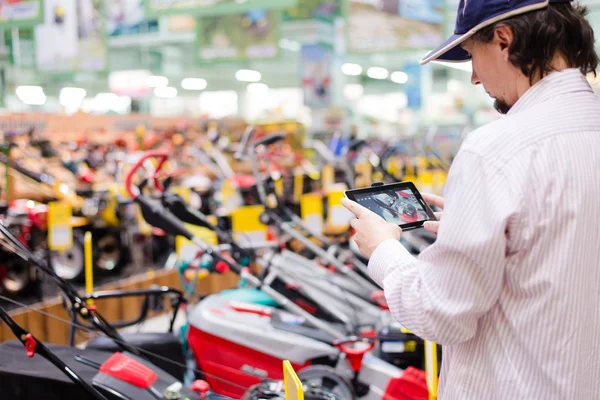 Young man choosing and photographing on the tablet pc computer lawnmowers in garden tools department of DIY shopping store background