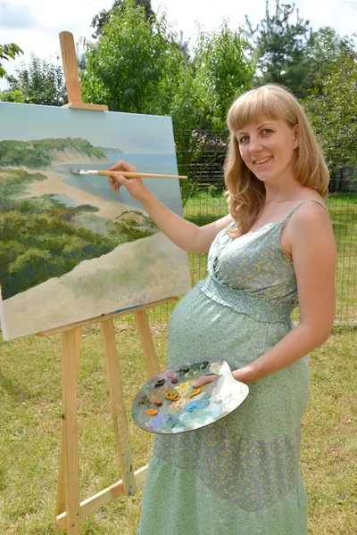 The young pregnant female artist draws a picture oil paints in t
