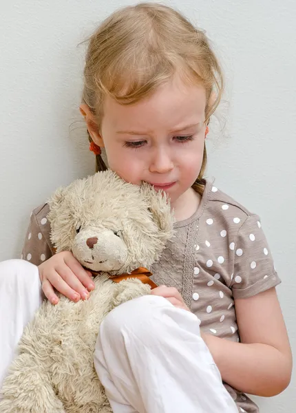 Sad lonely little girl sitting with teddy bear near the wall