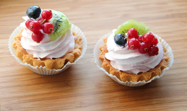 Two cute fancy cakes, cupcakes with creamy topping and berries -- red currants and blueberries on top against a wooden background
