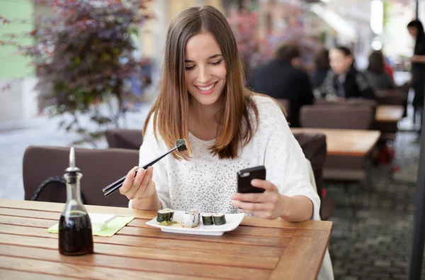 Happy young woman eating sushi in a restaurant and using mobile