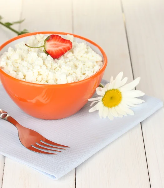 Cottage cheese with strawberry in orange bowl, fork and flower on blue napkin on white wooden table close-up