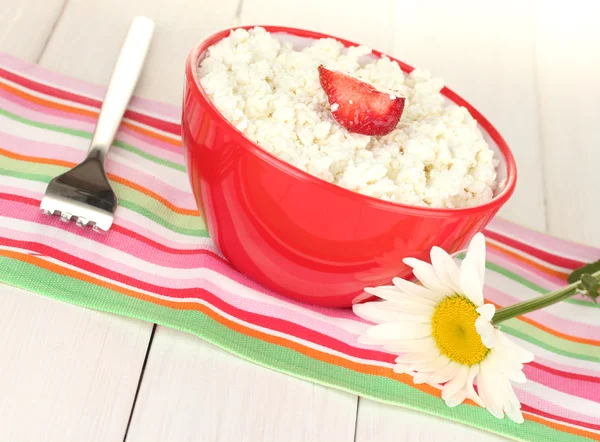 Cottage cheese with strawberry in red bowl, fork and flower on colorful napkin on white wooden table close-up