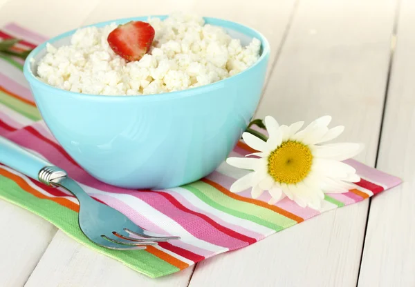Cottage cheese with strawberry in blue bowl, fork and flower on colorful napkin on white wooden table close-up