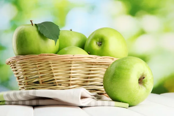 Ripe green apples with leaves in basket, on wooden table, on green backgrou