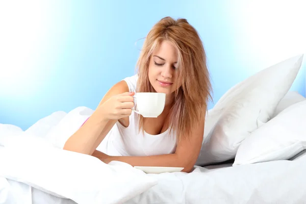 Young beautiful woman on bed with cup of coffee on blue background