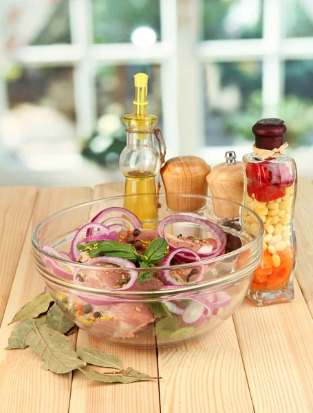 A large piece of pork marinated in bowl with herbs, spices and cooking oil on wooden table on window background